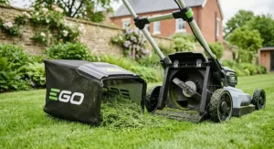 A high-detail, photorealistic 4K photograph focusing on the rear of the EGO cordless lawn mower with its grass collection box detached on the lawn, spilling a pile of fresh green grass clippings.