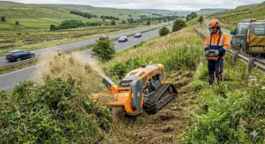 An industrial remote-controlled track mower cutting rough grass and invasive scrub on a severe highway embankment in the Yorkshire Dales.