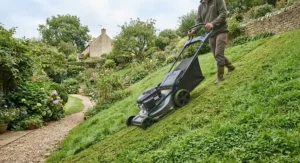Rear-wheel drive self-propelled lawn mower climbing a grassy bank in a residential garden.