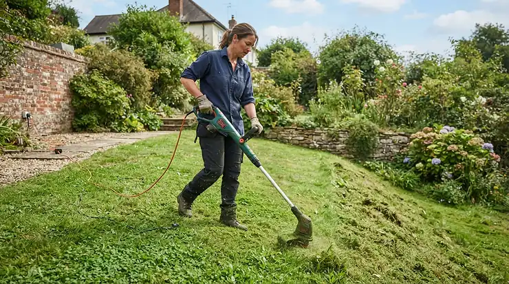Using a mains strimmer for large lawn areas, tackling overgrown grass along a steep garden bank in daylight.